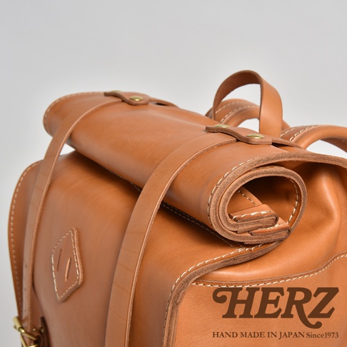 職人手作皮革露營登山用大容量後背包 R-19，棕色皮革材質，包身結構紮實，附有捲筒狀的固定帶和金屬扣件，縫線明顯，品牌標誌「HERZ」以燙金字體印製於包包一角。
