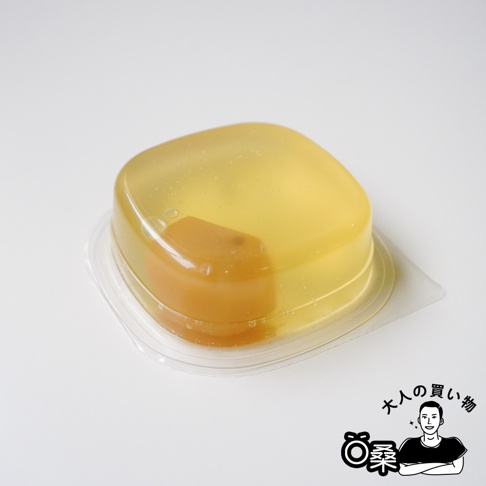 一個裝在透明塑膠盒裡的日式栗子蕨餅，呈現半透明的金黃色果凍質感，圓潤的栗子清晰可見地包覆在中間，背景為乾淨的白色，右下角配有大人的買い物標誌與人物插圖。