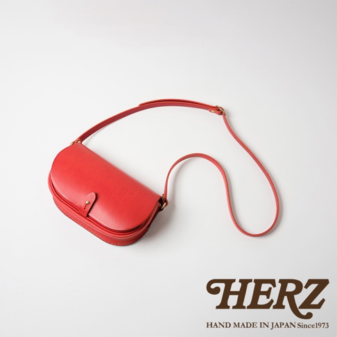 一個紅色的皮革迷你馬鞍包，具有彎曲的半月形輪廓，搭配可調節的細長肩帶和一個黃銅搭扣。