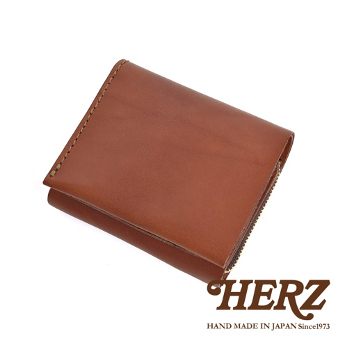 這是一款日本HERZ職人手工製作的棕色皮革短夾，呈現摺疊狀態，外觀簡約俐落，展現優質牛皮革的自然紋理與光澤，邊緣處有清晰的縫線細節，並在右下方印有品牌名稱與創立年份標誌。