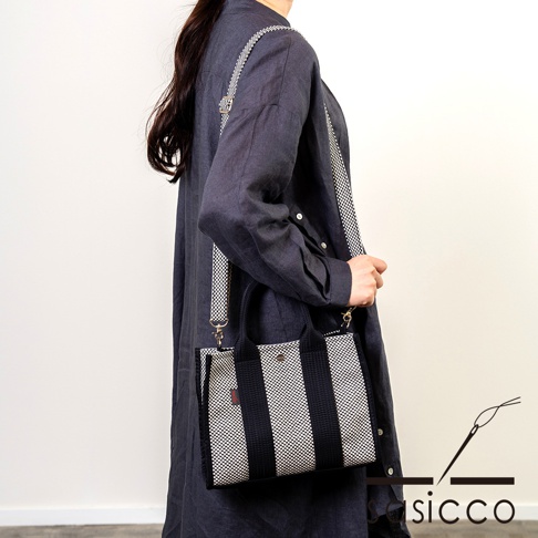 一名女性身穿深藍色長袖上衣，斜肩背著一款方形托特包。包包以三河木綿刺子織布料製成，有黑白格紋與深藍色條紋拼接設計，配有可拆卸的格紋肩帶。