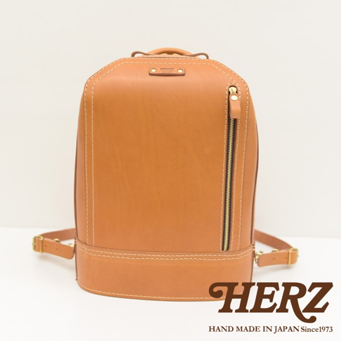 一個淺棕色皮革製的立體商務後背包，有著雙肩帶和一個垂直的拉鍊口袋，整體設計簡潔，展現職人手作質感。