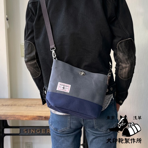 犬印純綿帆布雙色迷你側背包，採用灰色與深藍色拼接設計，配有深灰色可調式背帶。包包正面有一個印有品牌標誌的白色標籤。