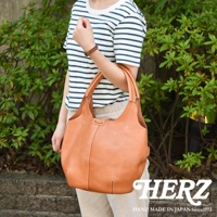 一位女士身著藍白條紋上衣和米色長褲，肩掛一個 tan 色皮革製的球形肩掛托特包，包包有著明顯的縫線和抽繩設計。