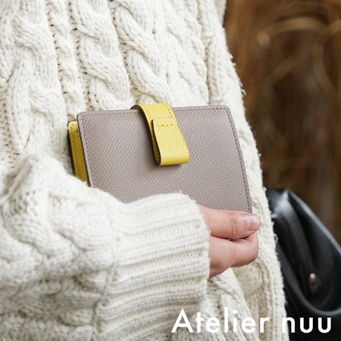 Atelier nuu 品牌 logo 顯示在圖片右下角，一位穿著奶油色針織毛衣的女士手裡拿著一個米灰色的皮革短夾，短夾上有一條亮黃色的皮帶做為裝飾。