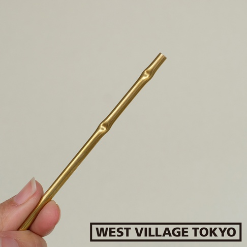 一根黃銅製成的細長線香架，表面呈現金屬光澤，並帶有仿竹節的紋路，一端為開放式管狀設計，由一隻手持握。