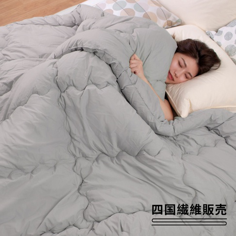 輕柔的淺灰色雙人冬被，表面有細緻的菱格車縫，一名女子被厚實的棉被包裹著，頭倚在米白色枕頭上。