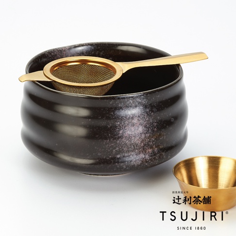 黑色陶瓷茶碗內置金色濾網，碗身帶有紋理和斑點，右側另有一個金色小碗。