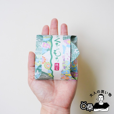 一隻手掌平托著一個包裝精緻的河內駿河屋和菓子，外層使用帶有傳統日式花草圖案的紙張包裹，中央腰封標示著抹茶口味的文字與符號，展現出雅緻的和風禮品設計。