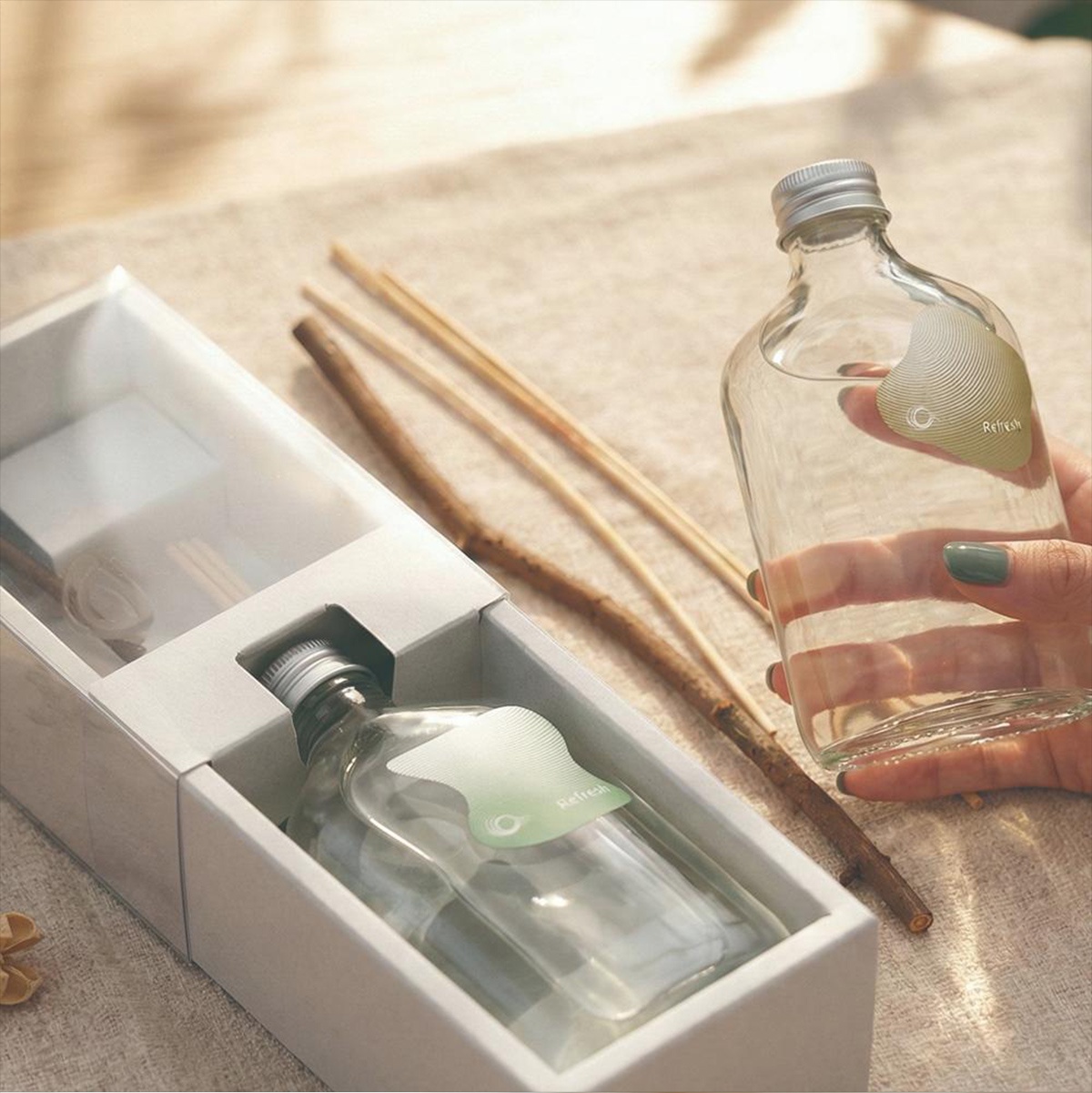 一瓶裝有透明香氛液的擴香瓶，瓶身上貼有綠色圓形標籤，上面印有「Refresh」字樣，瓶子由人手拿著，背景為兩根木質擴香棒和一個白色盒裝的擴香套組。