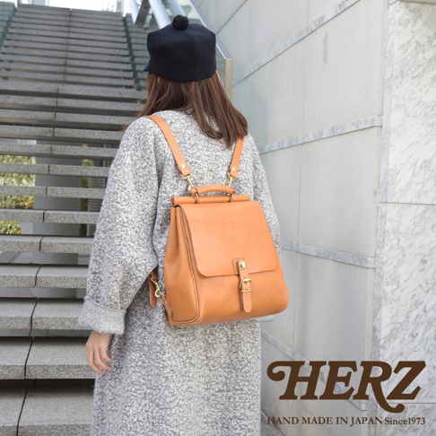 一位身穿灰色外套的女性背著一個棕褐色的皮革後背包，背包設計簡潔，有翻蓋和金屬扣環，散發著溫暖的復古氣息。