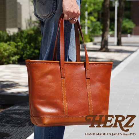 職人手作經典托特包 T-9 L，採用鞣製皮革製成，呈現溫潤的焦糖棕色。包款線條簡潔，提把與包身以同色系縫線細膩縫合，展現精緻工藝。