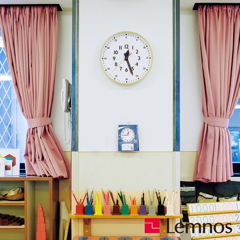 圓形白色蒙特梭利時鐘，黃色邊框，黑色數字時標，紅色時針與藍色分針，時鐘下方有彩色鉛筆，背景為粉色窗簾。