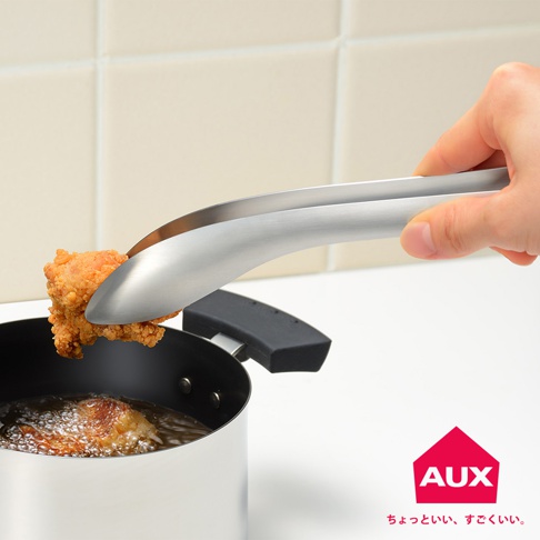 一隻手正使用銀色不鏽鋼曲型分菜夾夾起一塊炸雞，分菜夾的另一端放在一個銀色鍋子邊緣，鍋內有油和另一塊炸雞。