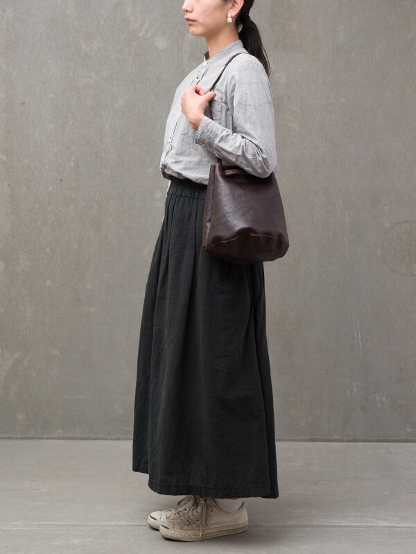 深棕色皮革抽繩水桶包，單肩背帶，包身圓潤，材質呈現自然紋理，搭配模特兒的灰色襯衫和黑色長裙。