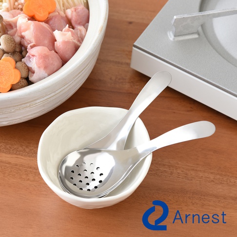 兩支不鏽鋼瀝水湯匙，其中一支湯匙帶有濾孔，置於白色小碗中，背景是裝有食材的鍋子和爐具。