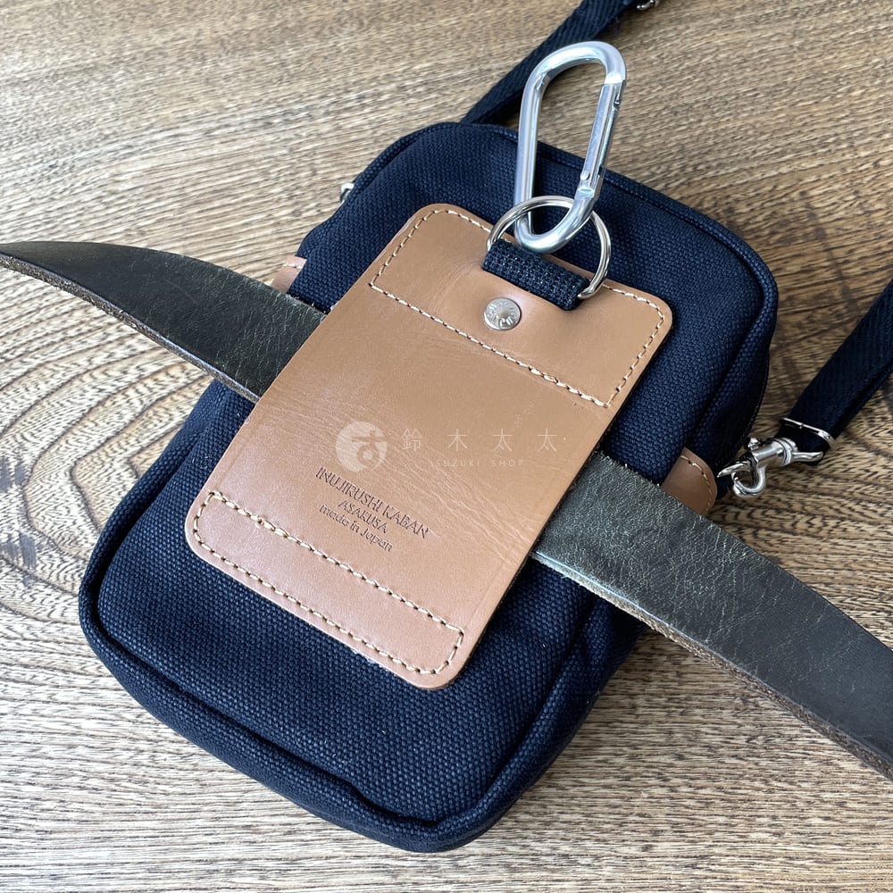 深藍色石洗帆布斜背手機包，配有棕色皮革吊牌，上面印有品牌標誌和「made in Japan」字樣，並有一個銀色登山扣。