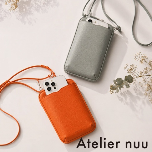 兩款 Atelier nuu 品牌皮革隨身手機包並排擺放，上方為灰綠色款式，下方為鮮豔橘色款式，兩者皆放入了智慧型手機，並配有細長斜背帶，背景為簡約淺色，裝飾有乾燥植物點綴。