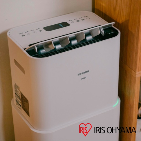 這台白色的 Iris Ohyama Pro 除濕機擺放在角落的木質家具旁，頂部設有觸控面板與顯示螢幕，面板上顯示著數字，機身前方印有品牌與系列名稱，底部邊緣處亮著綠色的指示燈。