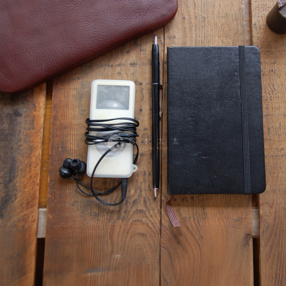 木紋背景上，一個白色 iPod，配有黑色耳機和纏繞的耳機線，旁邊是黑色鋼筆和黑色筆記本。左上方有一塊酒紅色皮革。