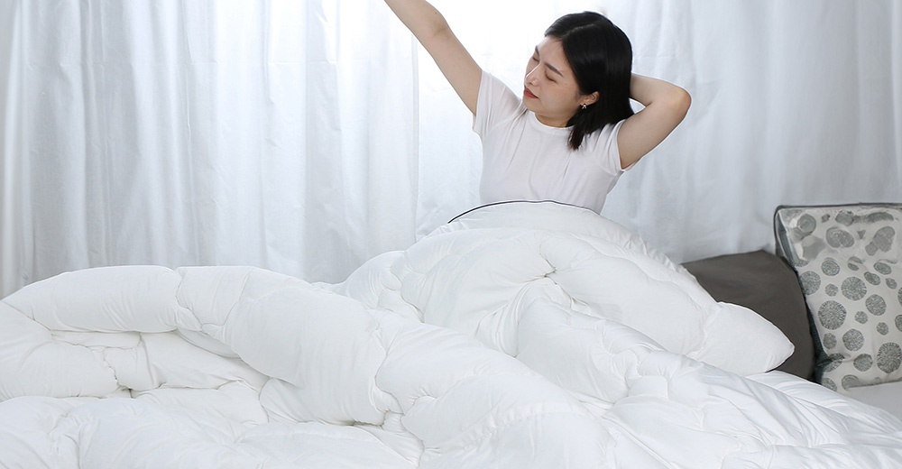 一位女子從被子裡伸出手臂打哈欠，背景是白色窗簾