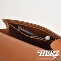 職人手作皮革翻蓋迷你後背包，焦糖棕色。內袋拉鍊開合，可見卡片與圓形物品。整體採用皮革材質，縫線清晰。