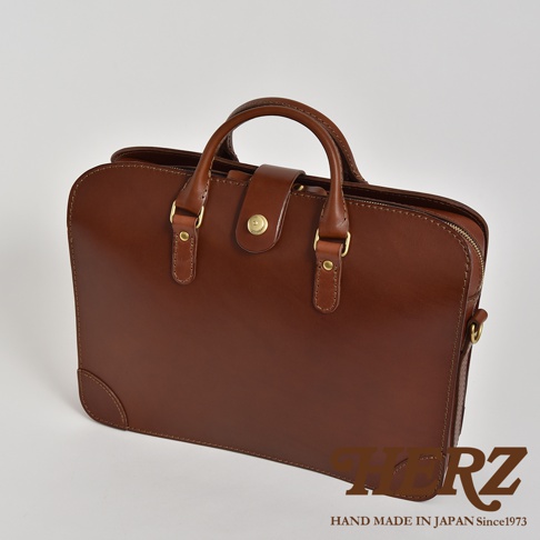 職人手作的棕色皮革商務公事包，設有三層收納空間，兩用設計，正面有皮帶扣與金色旋鈕，搭配圓潤提把與金色五金配件。