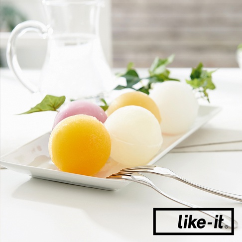 白色長方形盤子上擺放著四顆不同顏色的圓形冰球，分別是橘黃色、粉紅色、淡黃色和乳白色。背景有玻璃水瓶和綠葉裝飾，盤子旁放著兩支金屬叉子。