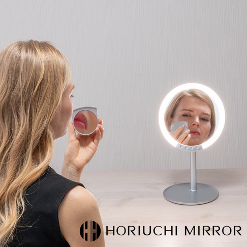 銀色金屬底座立式LED化妝鏡，圓形鏡面環繞著白色LED燈，鏡中映照著一位女士正在補妝。鏡前有一手持式迷你放大鏡，鏡頭聚焦於嘴唇。