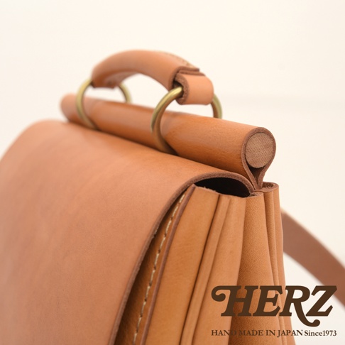 職人手作皮革屋瓦風後背包 R-15，以溫潤的淺棕色皮革製成，展現自然紋理與細緻車縫。包款頂部設有木質提把，以復古金屬環連接，整體設計簡約而有質感。