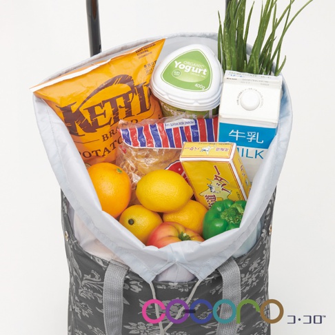 灰色印花提袋購物車內裝有數樣商品，包括橘子、檸檬、番茄、青椒、洋芋片、優格、牛奶盒、麵食包以及一小叢綠色植物。