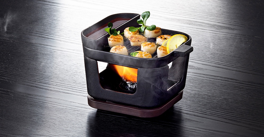 黑色鑄鐵桌上烤盤鍋爐組，烤盤上煎有數塊扇貝，並以檸檬片和綠葉裝飾，下方有火焰加熱。