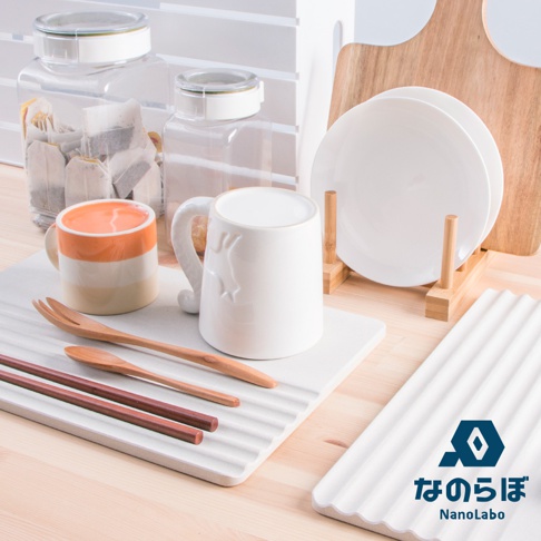 展示珪藻土瞬間吸水置物墊，墊上有木製叉子、湯匙和兩雙深棕色筷子。旁邊是橘白條紋馬克杯和白色浮雕馬克杯，後方有兩個裝有茶包的透明儲物罐，以及一個木製盤架上放著兩個白色盤子。