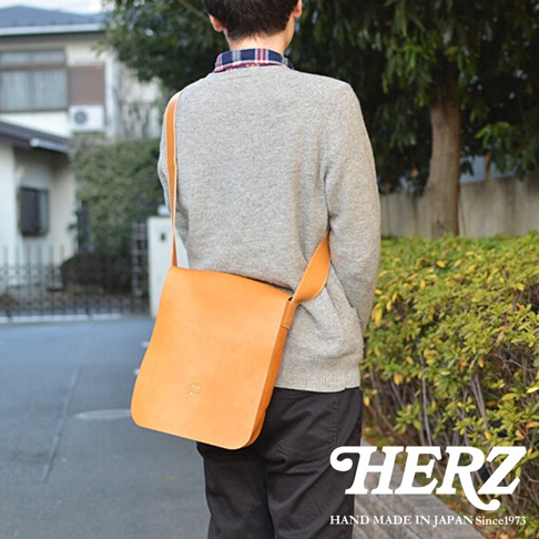 背對鏡頭的男子身穿灰色毛衣和格紋襯衫，側背一個淺棕色皮革斜背包，包上有品牌標誌。