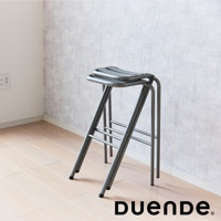 金屬材質的灰色高腳吧檯椅，椅面為弧形設計，椅腳為交叉結構，帶有橫向支撐桿，放在木質地板上，背景為淺色紋理牆面。