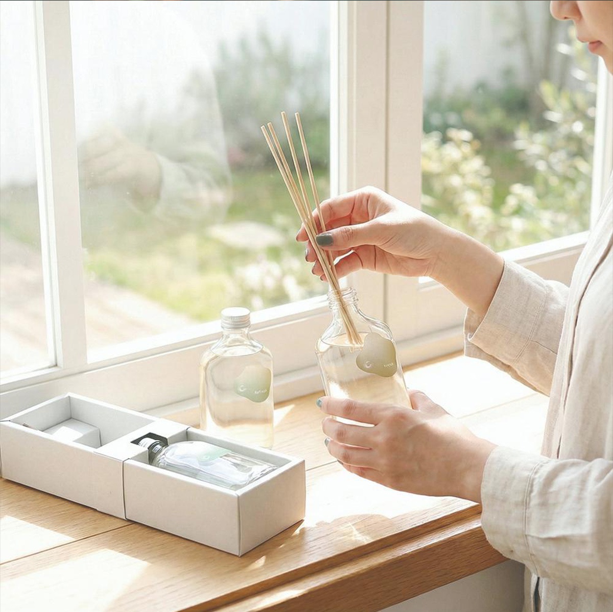 一名女子正將擴香竹插入玻璃瓶中，一旁另有一瓶擴香瓶與盒裝擴香瓶，背景是窗外綠意盎然的景象