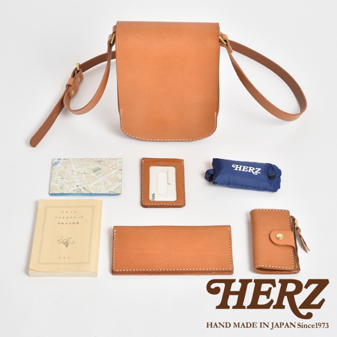 日本HERZ職人手作皮革翻蓋單肩包，呈現自然淺褐色皮革質感。包身上方為翻蓋直式設計，附有細長可調式肩帶。下方整齊擺放了隨身物品，包括地圖、皮革證件套、藍色HERZ收納袋、日文書籍、長夾及鑰匙包，整體構圖簡潔，展現濃厚的日式手工工藝風格。