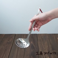 一只手正握著一把不鏽鋼萬用瀝水湯勺，勺面上佈滿圓形濾孔，勺柄末端有一個紅色掛繩。