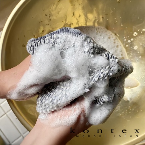 雙手正在搓揉一件含有藍色條紋、白色織物的洗澡巾，產生豐富泡沫，浸泡於金黃色盆中。