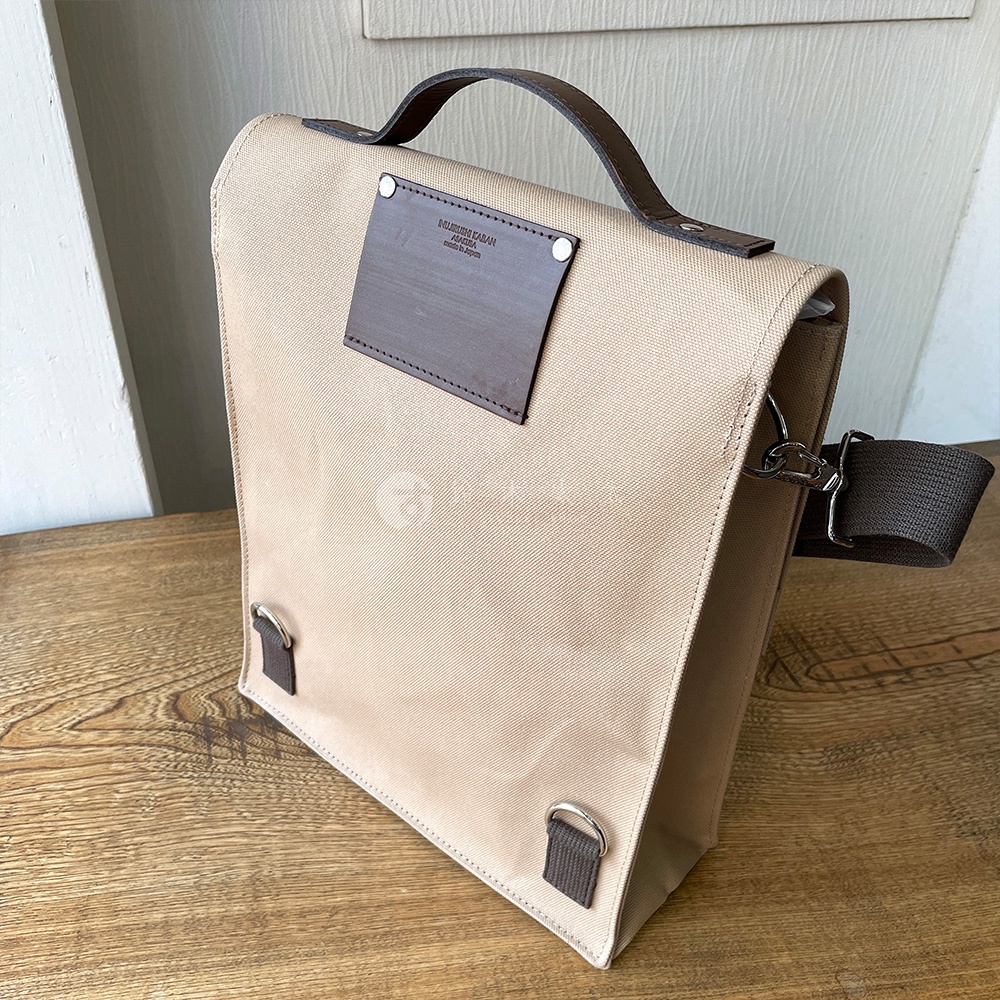 犬印純綿帆布三用直式側背包，卡其色帆布材質，搭配深棕色皮革提把與裝飾。包身有兩處金屬掛環，配有可拆卸的深棕色織帶。