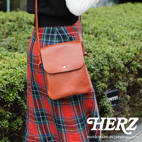 一位女性背著日本HERZ職人手作皮革隨行袋，包款呈現簡約的棕色皮革質感，搭配翻蓋設計與單顆金屬釦，背帶側邊帶有精緻的結飾細節，背景為戶外綠色灌木叢，右下角印有HERZ品牌字樣。