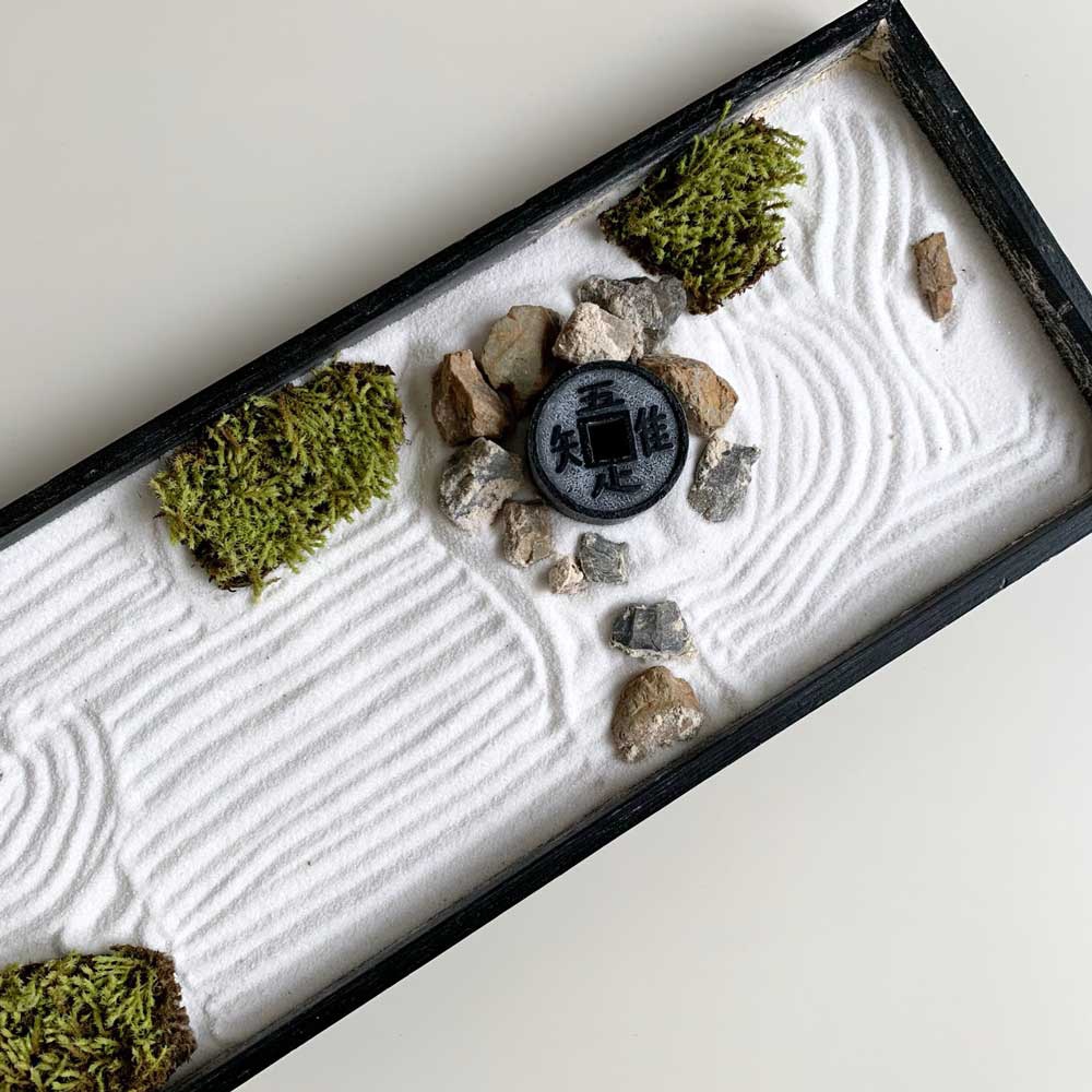 日式枯山水模型，黑色木框內盛有細緻的白色砂石，砂石上耙出波紋，點綴著數塊灰褐色石頭、翠綠色苔蘚塊，以及一枚帶有漢字的黑色圓形裝飾物。