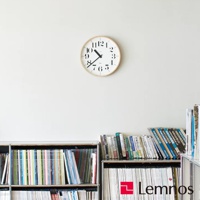 RIKI 時鐘(L)為圓形，白色錶盤搭配黑色數字和指針，外框為淺木色。時鐘懸掛於白色牆面上，下方擺放著兩個書架，架上陳列著各式書籍。