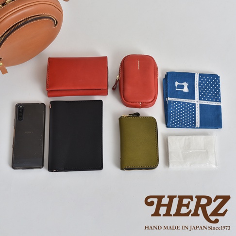 職人手作皮革小圓包 CK-19 S，展示了包包的收納能力。圖片中包含一個棕色皮革小圓包、紅色短夾、紅色零錢包、黑色長夾、綠色零錢包、手機、藍色印花手帕以及一包白色紙巾。