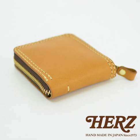 從側面拍攝的日本HERZ職人手作皮革全包式拉鍊短夾，採用淺棕色真皮材質，邊緣有明顯的車縫線細節，搭配黃銅色金屬拉鍊與皮革拉環，底部印有HERZ品牌商標與手作標語。
