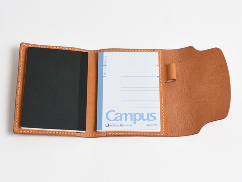 淺棕色皮革製成的A6記事本書套，內含一本黑色封面的筆記本和一本藍色「Campus」標誌的筆記本，書套邊緣有白色縫線，右側有一個皮革筆套。