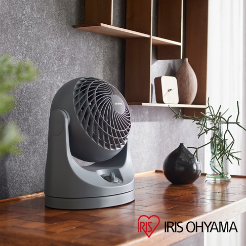 灰色的IRIS OHYAMA PCF-MKM15 空氣循環扇，具有可調節的傾斜角度和圓形網罩，放置在木質桌面上，背景為水泥牆面和置物架。