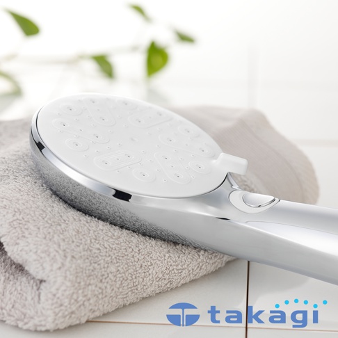這是一款 Takagi 真淨水水霧蓮蓬頭，金屬銀色握柄搭配白色圓形出水面板，平放在淺灰色毛巾上，面板邊緣設有切換開關，整體造型簡約俐落，背景為潔白的磁磚地與些許綠色植栽點綴。