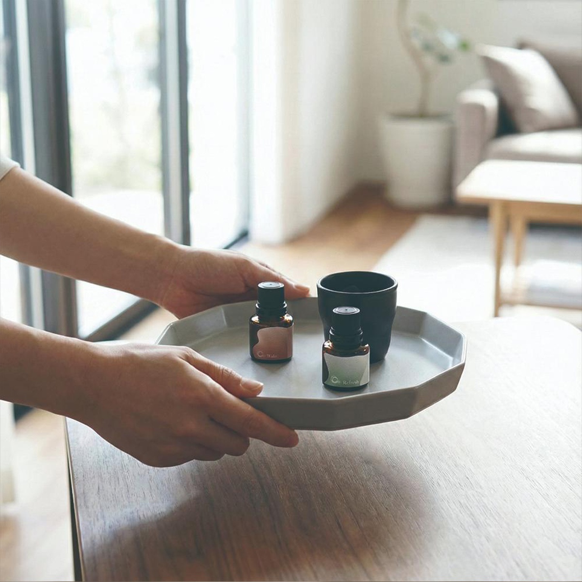 雙手捧著一個八邊形淺灰色盤子，上面放著兩個小型棕色精油瓶和一個黑色杯子。