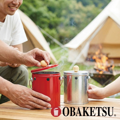 OBAKETSU 品牌標誌旁，兩隻手在一張木桌上，一隻手正在將一個木球放在紅色蚊香桶的蓋子上，另一個金屬罐子上也有一個木球，後面有營火和帳篷